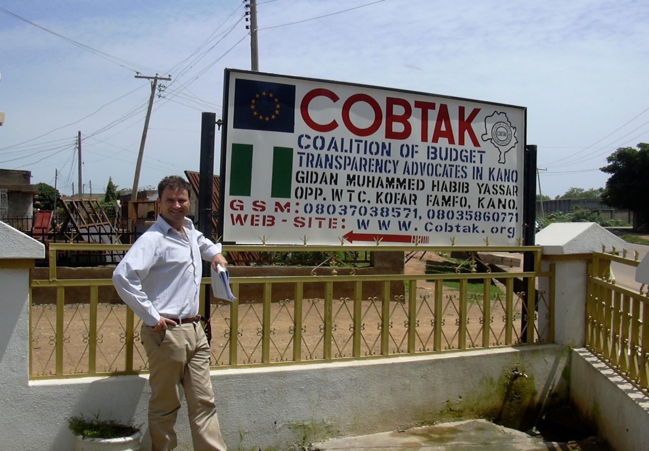 A visiting European Commission adviser beside the COBTAK office signpost, Kano.